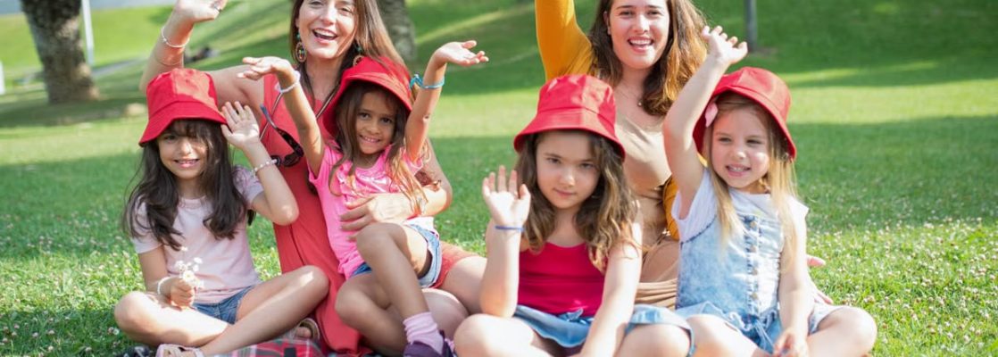 Two women and four girls enjoy a sunny picnic in Portugal, waving happily in the park.