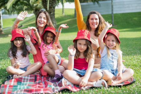 Two women and four girls enjoy a sunny picnic in Portugal, waving happily in the park.